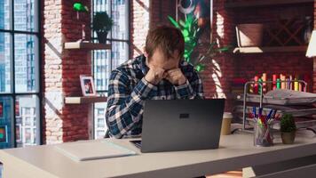 Tired man in living room office shrugging head, upset after having to answer too many emails on laptop. Exhausted freelancer feeling overworked while typing on device keyboard, camera A video