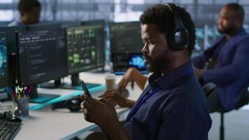 Admin in server room using mobile phone, relaxing after ensuring minimal downtime. Data center IT expert texting on smartphone, taking break from monitoring performance, camera A video