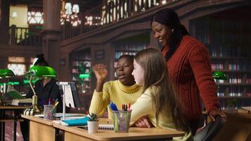 Elementary school pupils and tutor attend a live online course lesson from the library desk, connected via webcam to other classmates in a mentoring session. Virtual webinar discussion. Camera A. video