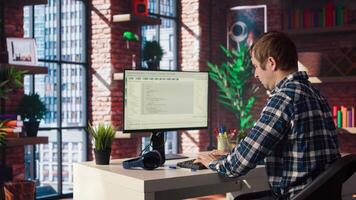 Man in home office stretches back while looking at UI on PC screen, feeling fatigued while executing programming code. IT developer feeling backpain, relieving pain, writing and testing code, camera A video