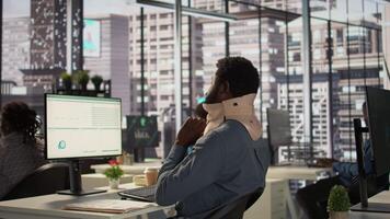 Hurt employee with a cervical collar working on a new business project, struggling to sit at the desk with his neck fracture. African american company associate tries to solve tasks. Camera B. video
