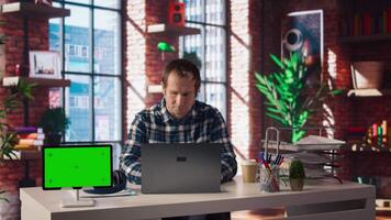 Green screen tablet next to employee completing spreadsheets on laptop and listening music. Chroma key device next to worker in home office wearing headphones editing documents, camera A video