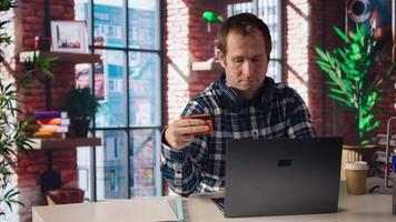 Happy man excited to do online shopping, typing debit card information on laptop in apartment. Cheerful person at desk using notebook, adding payment method on website, camera A video
