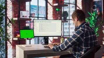 Man in home office looks at green screen tablet and computer, typing and executing programming code. Coding application used by IT developer writing and testing code using chroma key device, camera A video