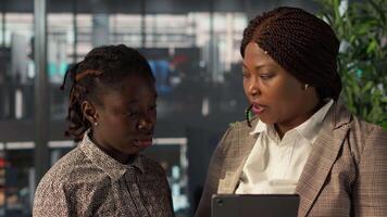 Black manager reviews company strategy with her female assistant in a high end corporate office, making key business decisions to support organizational growth. Discuss resource allocation. Camera A. video