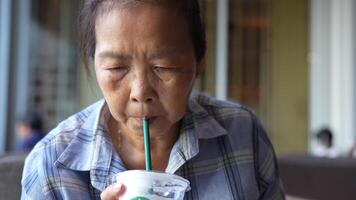 Elderly woman seated at a modern cafe table wearing a blue checkered shirt with a thoughtful expression. A drink and a pastry are placed in front of her surrounded by a relaxed ambience. video