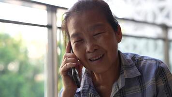 Senior woman using a smartphone near a large window with urban greenery outside, focusing intently on the screen. A scene blending modern technology with serene surroundings. video