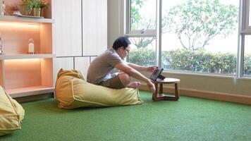 Casual indoor workspace scene featuring a man sitting on a beanbag chair, using a tablet on a small wooden table. Warm lighting and a green carpet create a relaxed and cozy atmosphere. video