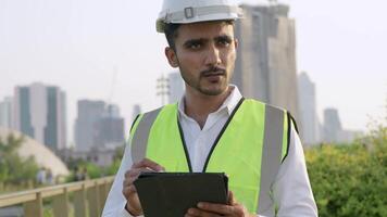 Engineer in a white helmet and high-visibility vest holding a tablet, gesturing confidently outdoors. Urban skyline and construction cranes in the background symbolize modern infrastructure. video