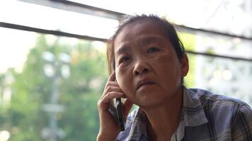Senior woman using a smartphone near a large window with urban greenery outside, focusing intently on the screen. A scene blending modern technology with serene surroundings. video