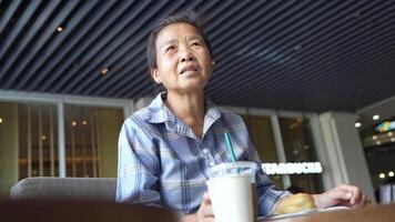 Elderly woman seated at a modern cafe table wearing a blue checkered shirt with a thoughtful expression. A drink and a pastry are placed in front of her surrounded by a relaxed ambience. video