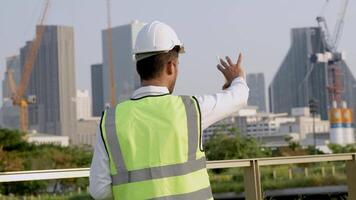 Engineer in a white helmet and high-visibility vest holding a tablet, gesturing confidently outdoors. Urban skyline and construction cranes in the background symbolize modern infrastructure. video