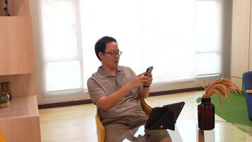 Man seated at a modern glass table in a bright, minimalist living space, holding a camera and interacting with a tablet. Shelving and yellow chairs add a cozy touch to the aesthetic. video