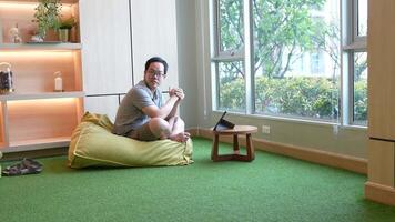 Casual indoor workspace scene featuring a man sitting on a beanbag chair, using a tablet on a small wooden table. Warm lighting and a green carpet create a relaxed and cozy atmosphere. video