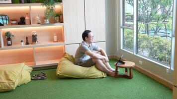 Casual indoor workspace scene featuring a man sitting on a beanbag chair, using a tablet on a small wooden table. Warm lighting and a green carpet create a relaxed and cozy atmosphere. video