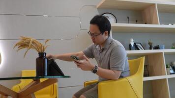 Man seated at a modern glass table in a bright, minimalist living space, holding a camera and interacting with a tablet. Shelving and yellow chairs add a cozy touch to the aesthetic. video