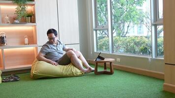 Casual indoor workspace scene featuring a man sitting on a beanbag chair, using a tablet on a small wooden table. Warm lighting and a green carpet create a relaxed and cozy atmosphere. video