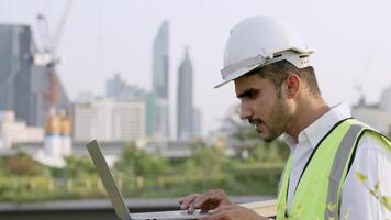 Engineer in a white helmet and high-visibility vest holding a tablet, gesturing confidently outdoors. Urban skyline and construction cranes in the background symbolize modern infrastructure. video