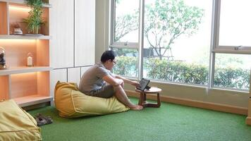 Casual indoor workspace scene featuring a man sitting on a beanbag chair, using a tablet on a small wooden table. Warm lighting and a green carpet create a relaxed and cozy atmosphere. video