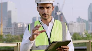 Engineer in a white helmet and high-visibility vest holding a tablet, gesturing confidently outdoors. Urban skyline and construction cranes in the background symbolize modern infrastructure. video