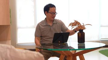Man seated at a modern glass table in a bright, minimalist living space, holding a camera and interacting with a tablet. Shelving and yellow chairs add a cozy touch to the aesthetic. video