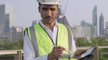 Engineer in a white helmet and high-visibility vest holding a tablet, gesturing confidently outdoors. Urban skyline and construction cranes in the background symbolize modern infrastructure. video