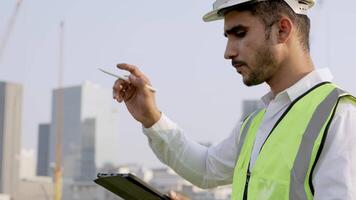Engineer in a white helmet and high-visibility vest holding a tablet, gesturing confidently outdoors. Urban skyline and construction cranes in the background symbolize modern infrastructure. video
