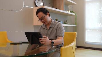 Man seated at a modern glass table in a bright, minimalist living space, holding a camera and interacting with a tablet. Shelving and yellow chairs add a cozy touch to the aesthetic. video