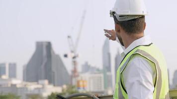 Engineer in a white helmet and high-visibility vest holding a tablet, gesturing confidently outdoors. Urban skyline and construction cranes in the background symbolize modern infrastructure. video