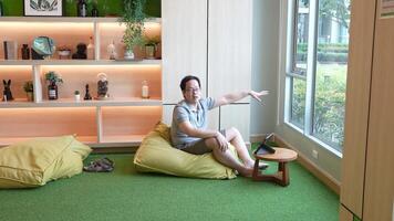 Casual indoor workspace scene featuring a man sitting on a beanbag chair, using a tablet on a small wooden table. Warm lighting and a green carpet create a relaxed and cozy atmosphere. video