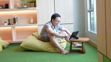 Casual indoor workspace scene featuring a man sitting on a beanbag chair, using a tablet on a small wooden table. Warm lighting and a green carpet create a relaxed and cozy atmosphere. video