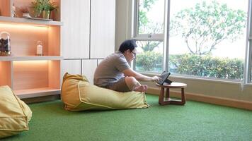 décontractée intérieur espace de travail scène avec une homme séance sur une pouf chaise, en utilisant une tablette sur une petit en bois tableau. chaud éclairage et une vert tapis créer une détendu et confortable atmosphère. video