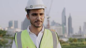 Engineer in a white helmet and high-visibility vest holding a tablet, gesturing confidently outdoors. Urban skyline and construction cranes in the background symbolize modern infrastructure. video