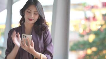 Elegant woman in a purple blazer using her smartphone, gracefully waving in a modern outdoor setting. A blend of style, technology, and sophistication with a warm and cheerful expression. video