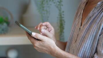 Senior woman using a smartphone near a large window with urban greenery outside, focusing intently on the screen. A scene blending modern technology with serene surroundings. video