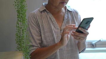 Senior woman using a smartphone near a large window with urban greenery outside, focusing intently on the screen. A scene blending modern technology with serene surroundings. video