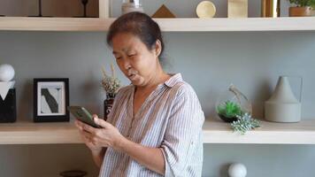 Senior woman using a smartphone near a large window with urban greenery outside, focusing intently on the screen. A scene blending modern technology with serene surroundings. video