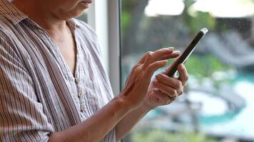 Senior woman using a smartphone near a large window with urban greenery outside, focusing intently on the screen. A scene blending modern technology with serene surroundings. video