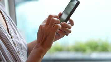 Senior woman using a smartphone near a large window with urban greenery outside, focusing intently on the screen. A scene blending modern technology with serene surroundings. video