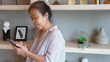 Senior woman using a smartphone near a large window with urban greenery outside, focusing intently on the screen. A scene blending modern technology with serene surroundings. video