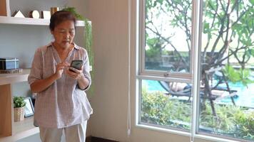 Senior woman using a smartphone near a large window with urban greenery outside, focusing intently on the screen. A scene blending modern technology with serene surroundings. video