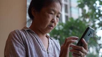Senior woman using a smartphone near a large window with urban greenery outside, focusing intently on the screen. A scene blending modern technology with serene surroundings. video