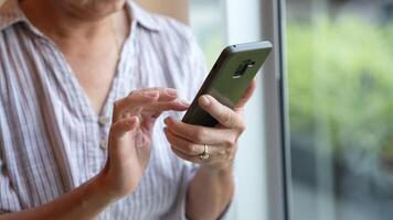 Senior woman using a smartphone near a large window with urban greenery outside, focusing intently on the screen. A scene blending modern technology with serene surroundings. video