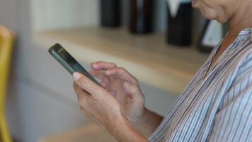 Senior woman using a smartphone near a large window with urban greenery outside, focusing intently on the screen. A scene blending modern technology with serene surroundings. video