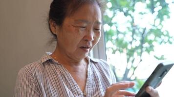 Senior woman using a smartphone near a large window with urban greenery outside, focusing intently on the screen. A scene blending modern technology with serene surroundings. video
