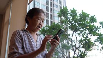 Senior woman using a smartphone near a large window with urban greenery outside, focusing intently on the screen. A scene blending modern technology with serene surroundings. video