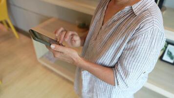Senior woman using a smartphone near a large window with urban greenery outside, focusing intently on the screen. A scene blending modern technology with serene surroundings. video