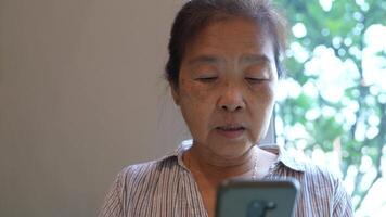 Senior woman using a smartphone near a large window with urban greenery outside, focusing intently on the screen. A scene blending modern technology with serene surroundings. video