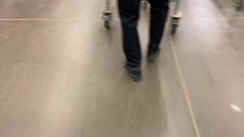The feet of a shopper can be seen walking in the supermarket aisle while pushing a cart, showcasing a bustling shopping environment filled with numerous other customers wandering around video