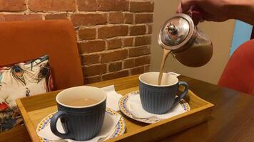 A hand pours warm tea from a silver pot into stylish blue cups, set on a wooden tray amidst a charming caf atmosphere video